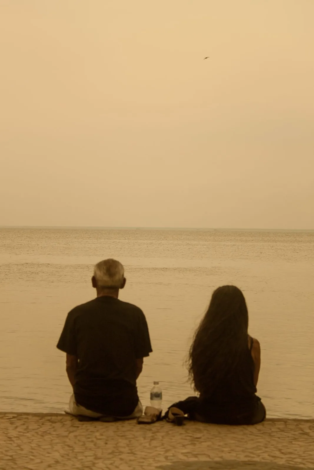 Duas pessoas sentadas de frente para o mar, quase escuro